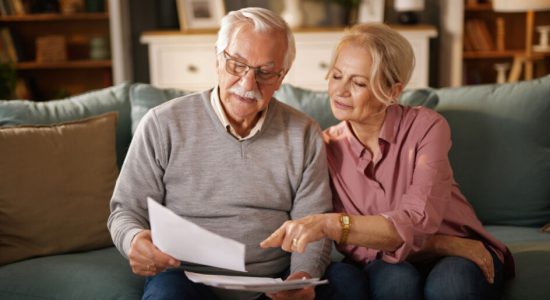 Elderly couple reviewing life insurance policy documents, planning their future finances together on the sofa